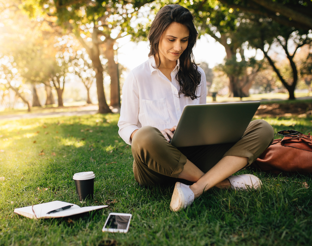 Young businesswoman working on her notebook in park. Freelancer female sitting on grassy lawn using laptop computer with her diary, mobile phone and coffee at her side on grass.